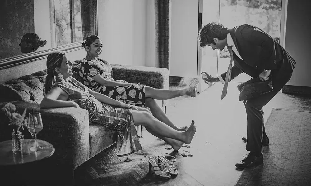 Ein Hochzeitsgast sprüht lachend die Füße einer Brautjungfer mit Deodorant ein.