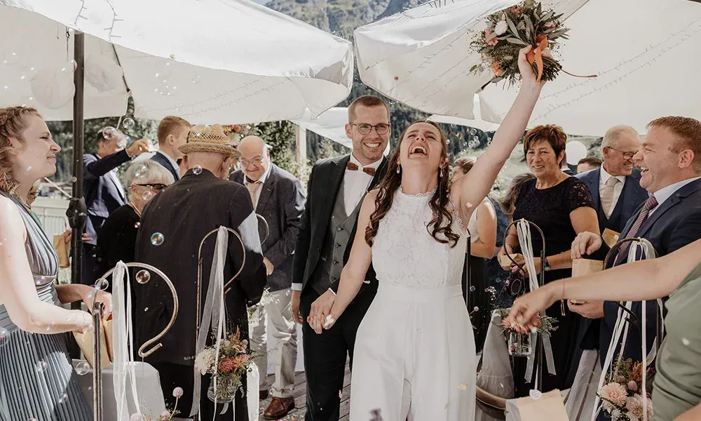 Die Braut reißt jubelnd den Strauß in die Luft, umgeben von feiernden Gästen.