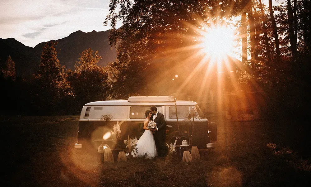 Brautpaar küsst sich im Sonnenuntergang vor einem Retro-Bus inmitten der Natur.
