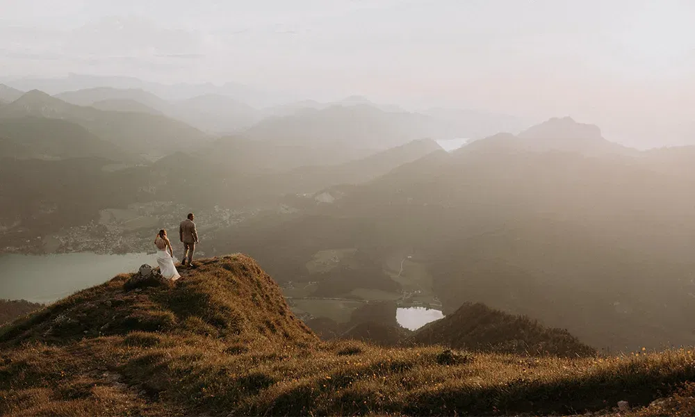 Hochzeitspaar auf Berggipfel mit atemberaubender Aussicht und weichem Abendlicht.