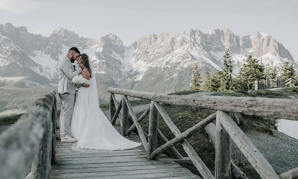 Brautpaar auf Holzbrücke mit Blick auf verschneite Bergkulisse, verliebt aneinander gelehnt.