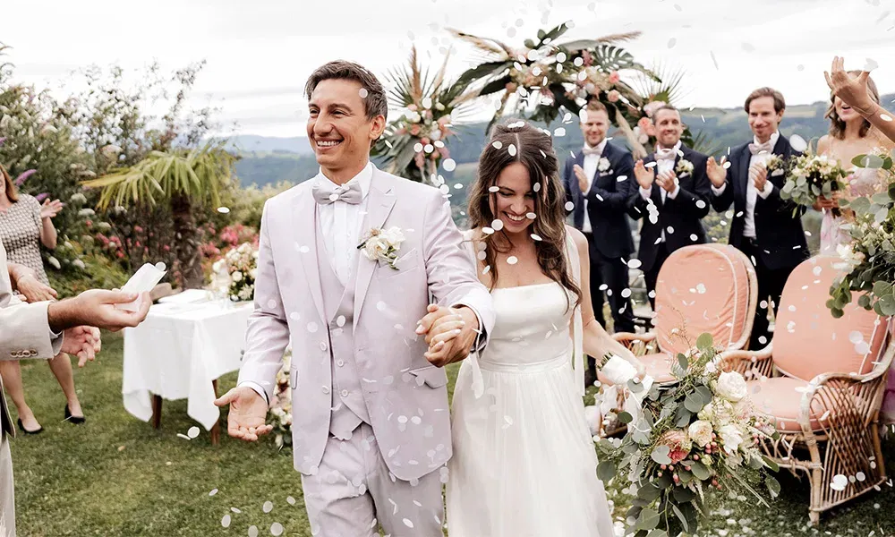 Glücklicher Bräutigam und strahlende Braut verlassen Hand in Hand die freie Trauung unter einem Konfettiregen.