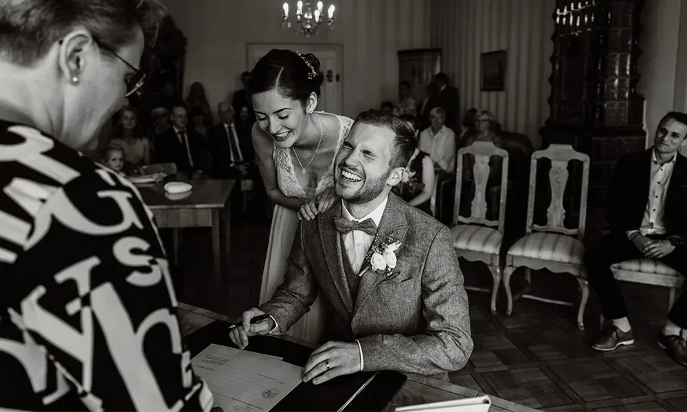 Bräutigam lacht herzhaft beim Unterschreiben der Heiratsurkunde, Braut steht liebevoll hinter ihm.