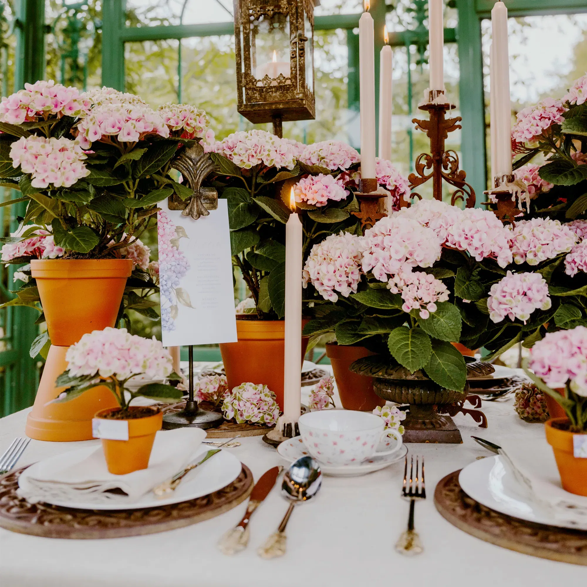 Liebevoll gedeckter Hochzeitstisch mit rosa Hortensien in Tontöpfen, Vintage-Geschirr und Kerzenleuchtern im Grünen.