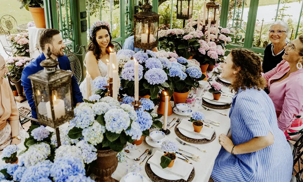 Brautpaar mit Gästen an festlich dekorierter DIY-Tafel voller Topf-Hortensien in Blau und Rosa, umgeben von Laternen und Kerzenschein.