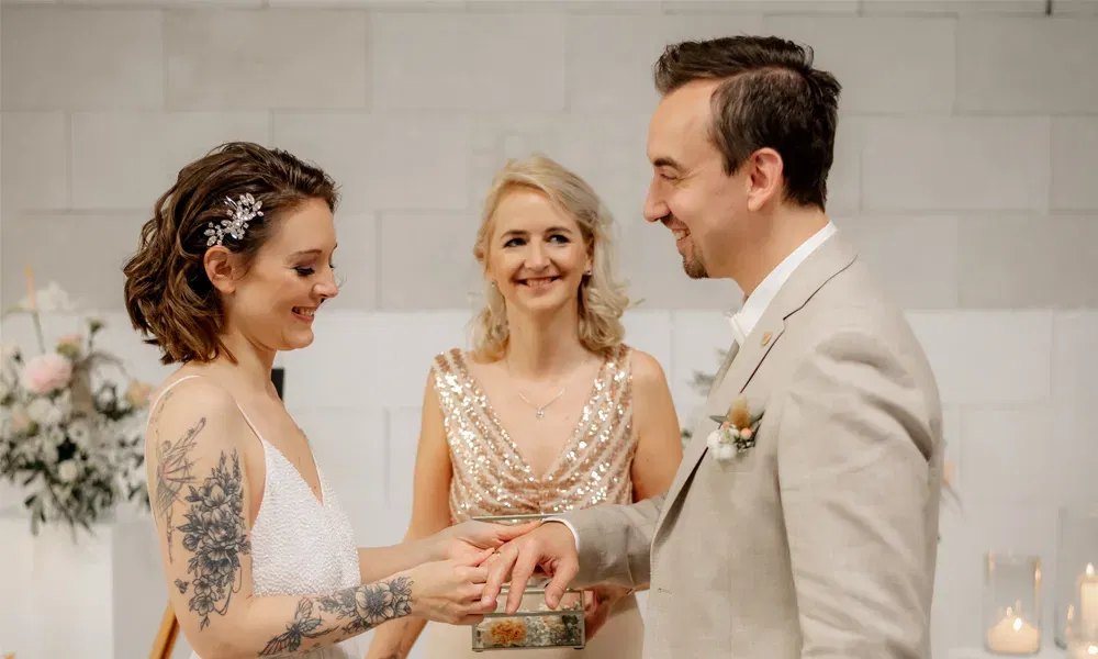 Freie Traurednerin beobachtet mit einem warmen Lächeln, wie Braut und Bräutigam sich bei der Zeremonie gegenseitig die Ringe anstecken.