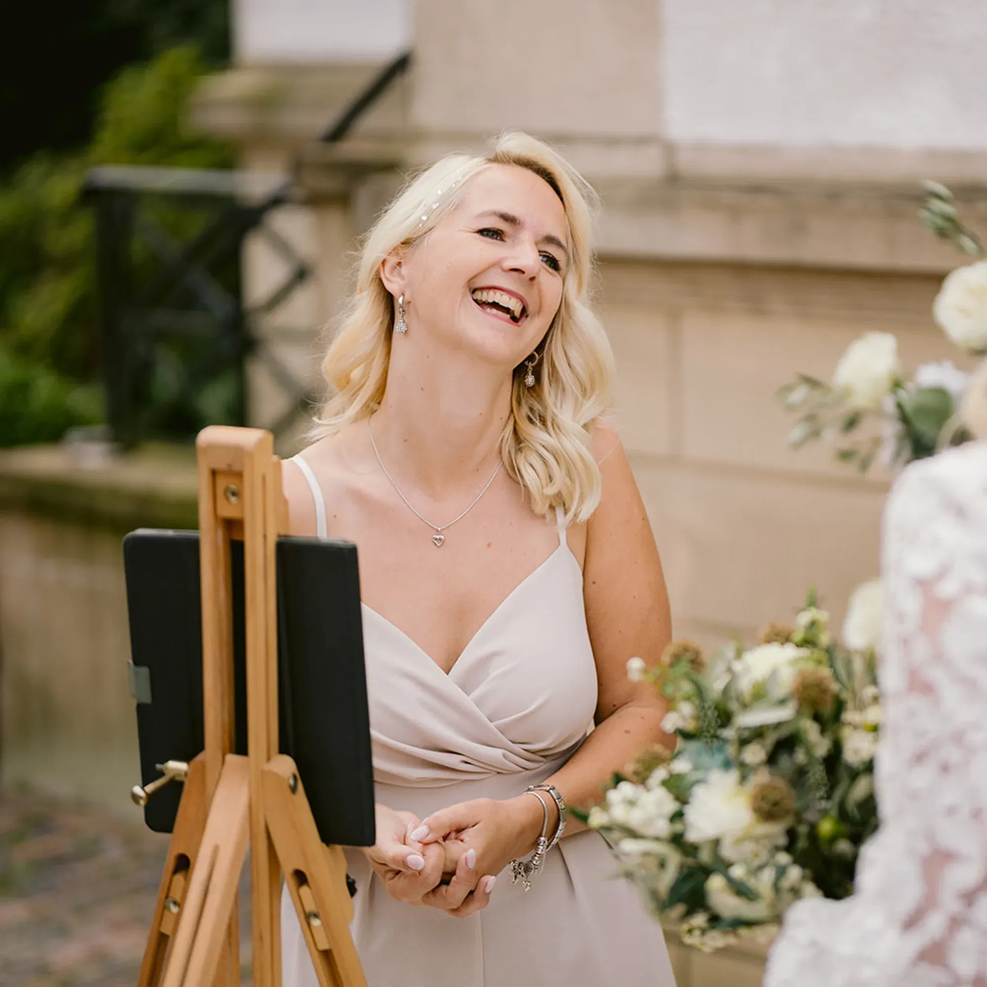 Hochzeitsrednerin im cremefarbenen Kleid lächelt herzlich während einer Zeremonie unter freiem Himmel.