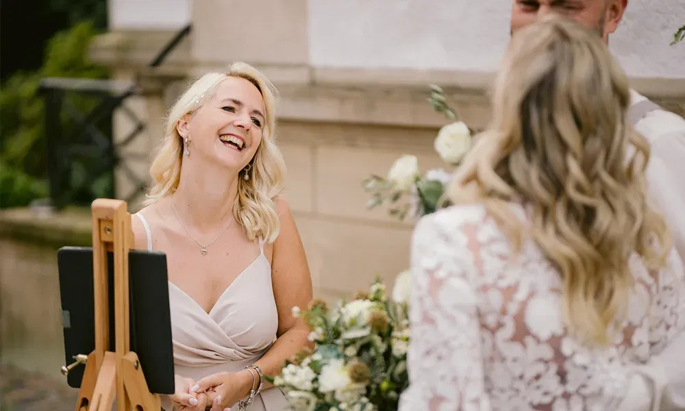 Hochzeitsrednerin im hellen Kleid lächelt offen während einer Zeremonie unter freiem Himmel – Braut und Bräutigam im Vordergrund.