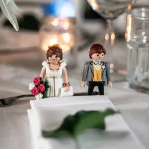 Nahaufnahme eines Hochzeitstischs mit Playmobil-Figuren im Brautkleid und Anzug, umgeben von Kerzenschein und Gläsern.