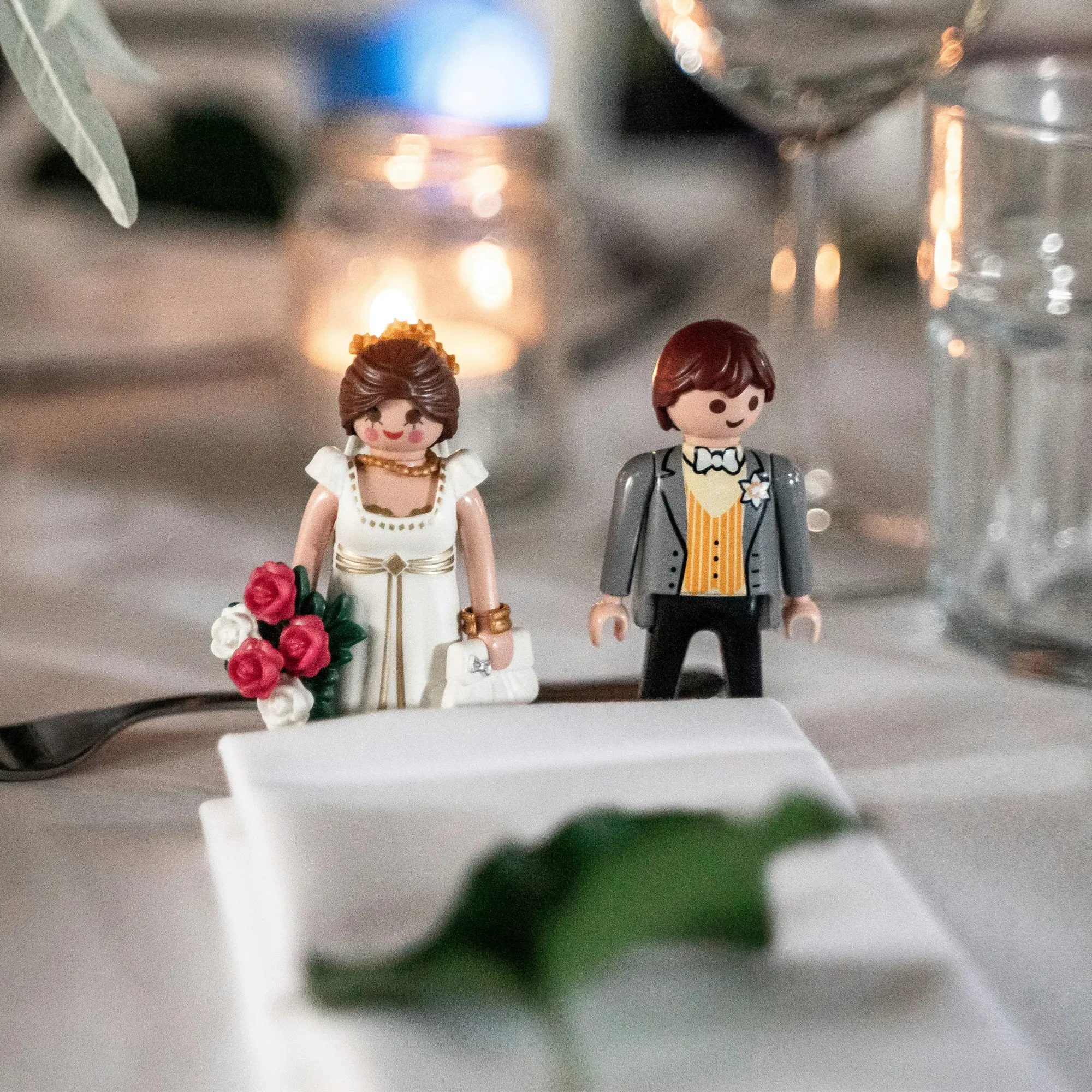 Nahaufnahme eines Hochzeitstischs mit Playmobil-Figuren im Brautkleid und Anzug, umgeben von Kerzenschein und Gläsern.