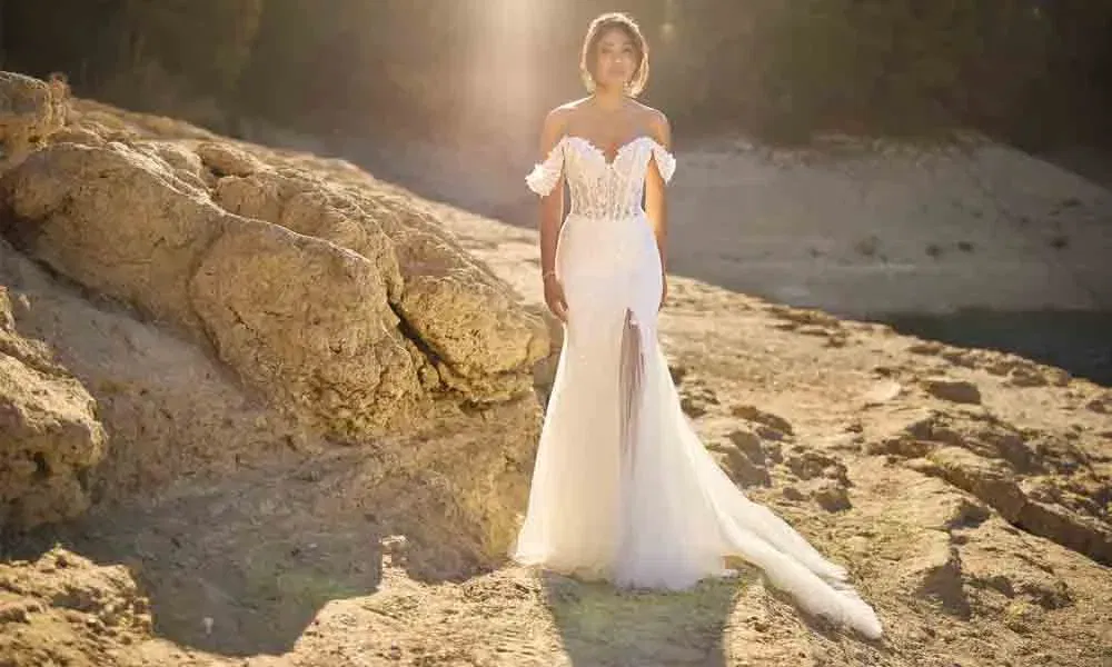 Hochzeitskleid mit Spitzenoberteil und Carmen-Ausschnitt, getragen von einer Braut auf einem sonnigen Felsplateau vor bewaldetem Hintergrund.