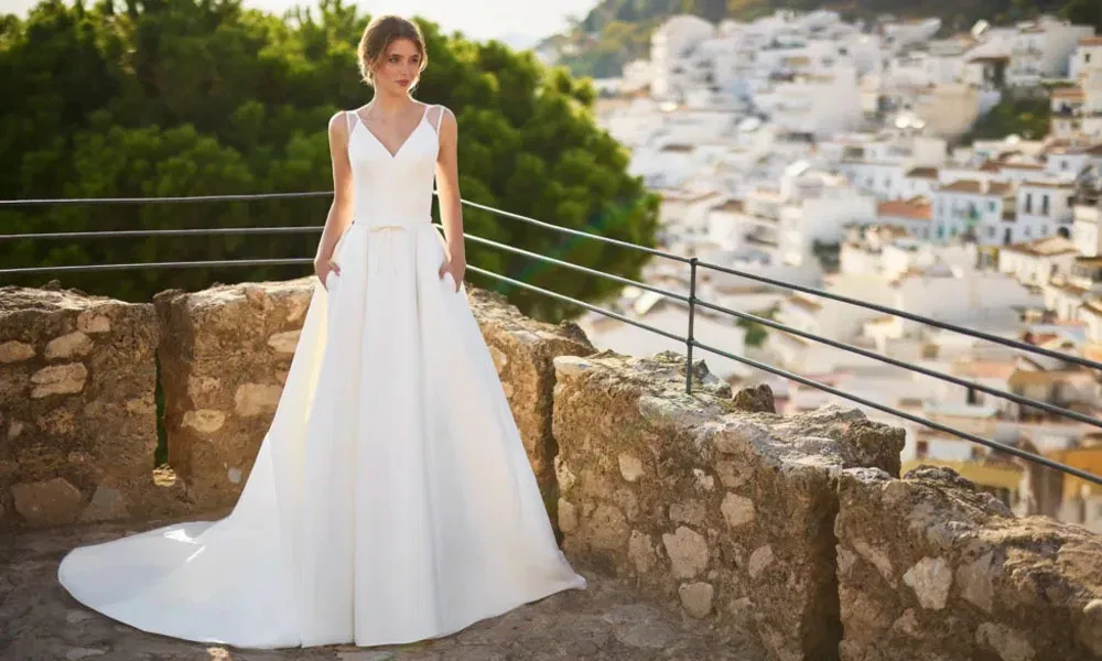 Elegante Braut im minimalistischen Hochzeitskleid mit Schleppe steht auf einer Burgterrasse mit Blick auf weiße Häuser und grüne Hügel.