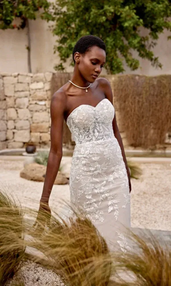 Elegante Braut im schulterfreien Hochzeitskleid mit floraler Spitze, entspannt im rustikalen Gartenambiente posierend.