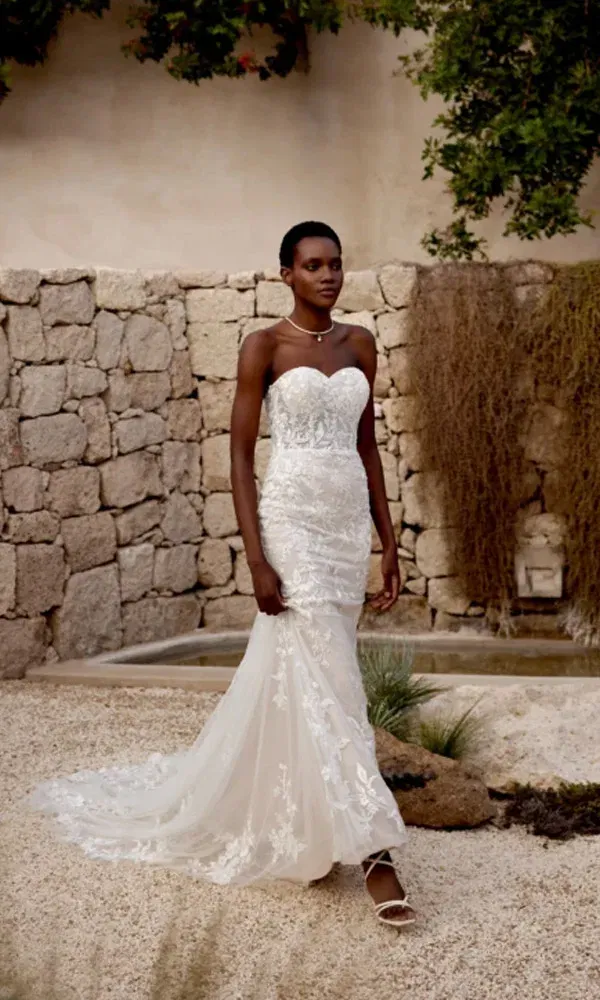 Hochzeitskleid mit Spitze, trägerlos und elegant – Braut läuft barfuß durch hellen Garten mit Naturmauer.