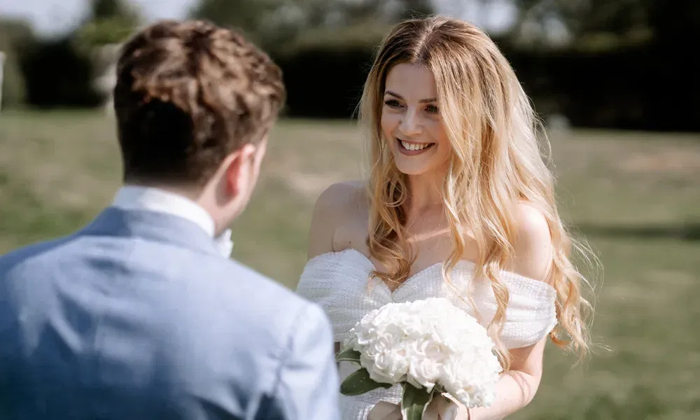 Romantischer Moment unter freiem Himmel: Die Braut im Off-Shoulder-Kleid blickt ihrem Partner lächelnd entgegen und hält einen weißen Blumenstrauß.