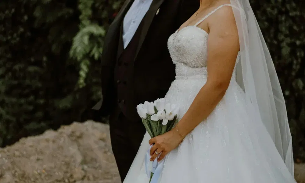 Detailaufnahme eines Hochzeitspaares: Die Braut trägt ein glitzerndes Trägerkleid mit Schleier und Blumenstrauß, der Bräutigam trägt einen schwarzen Anzug.