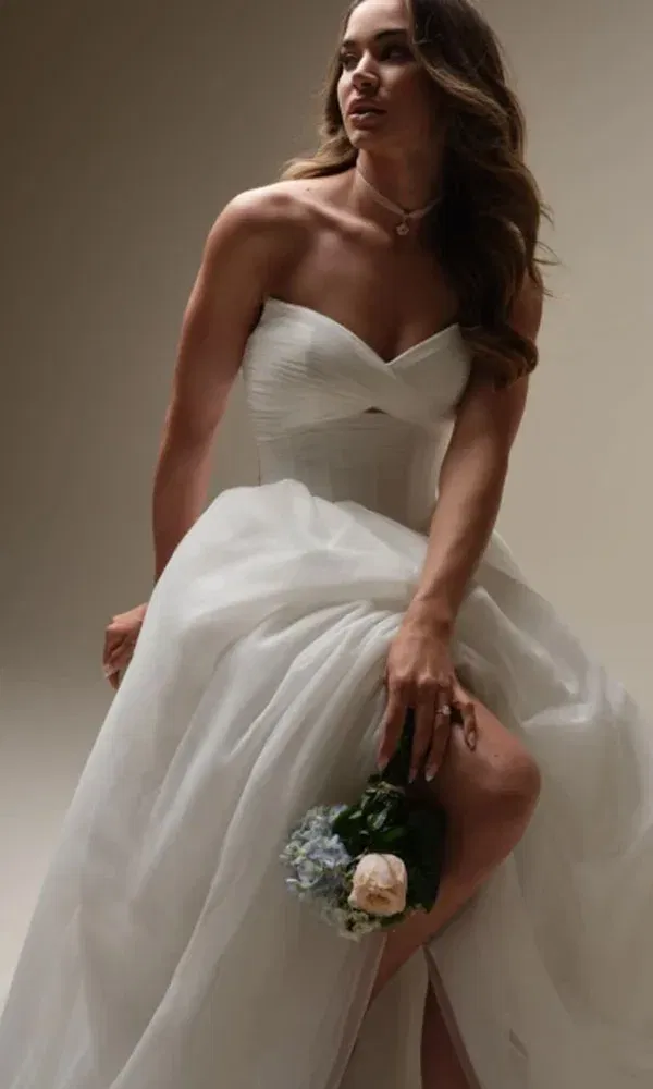 Braut mit wallendem Tüllkleid und romantischem Ausschnitt posiert elegant mit Blumen auf dem Schoß.