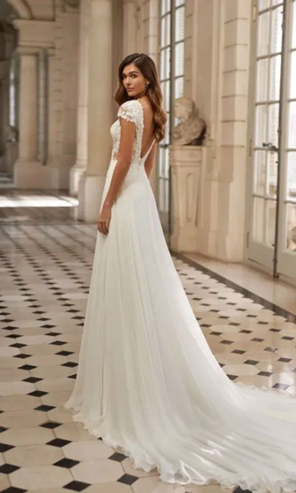 Elegantes Brautkleid mit tiefer Rückansicht, Spitzenärmeln und zartem Chiffonrock – Braut blickt über die Schulter in einem Schlossinterieur.