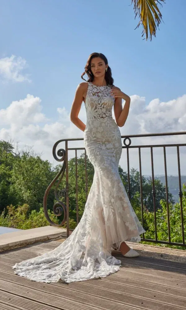Braut in figurbetontem Kleid mit hoher Illusions-Halslinie und floralen Applikationen auf Holzdeck mit Aussicht.