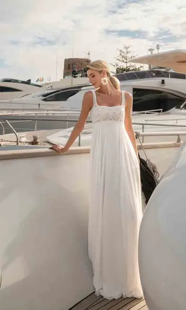 Elegantes Empire-Kleid mit Spitzenträgern, getragen von einer Braut auf einem Bootssteg mit luxuriösen Yachten im Hintergrund.
