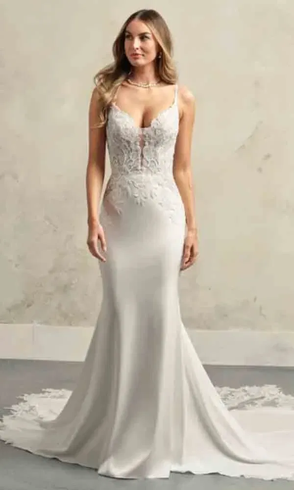 Moderne Braut mit figurbetontem Brautkleid mit tiefem Ausschnitt und floraler Spitze auf schlichtem Untergrund.