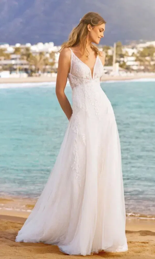 Sommerliche Strandhochzeit mit einer Braut im leichten Tüllkleid und tiefem Ausschnitt, die auf den Ozean blickt.