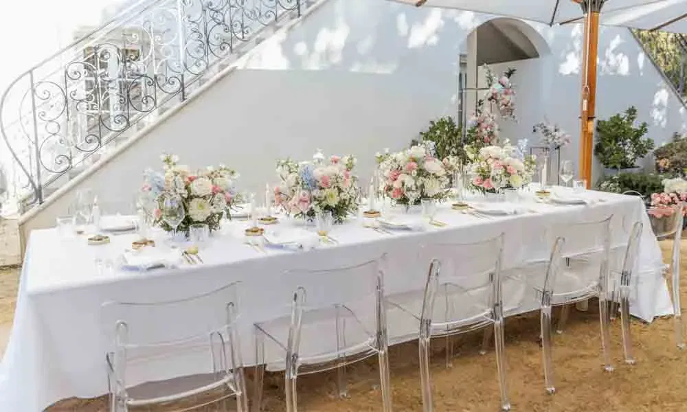 Romantisch gedeckter Tisch im Garten mit opulenter Blumendeko in Rosa, Creme und Blau, umgeben von Acrylstühlen und Sonnenschirm.