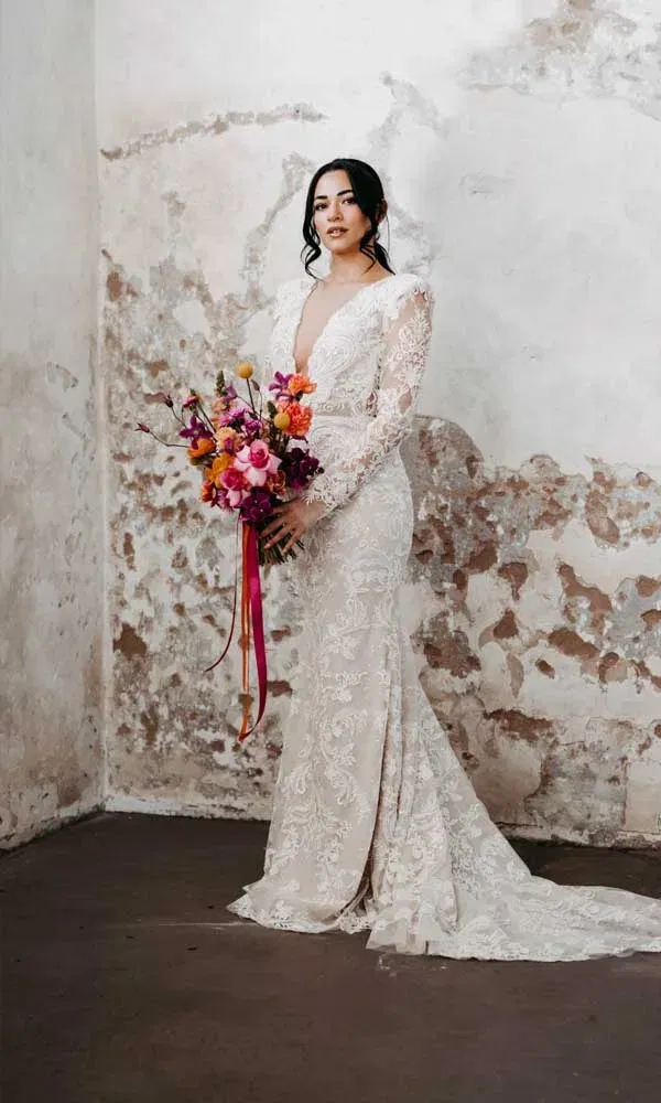 Stilvolle Braut im figurbetonten Spitzenkleid mit langen Ärmeln, hält bunte Blumen vor rauer, heller Wandkulisse