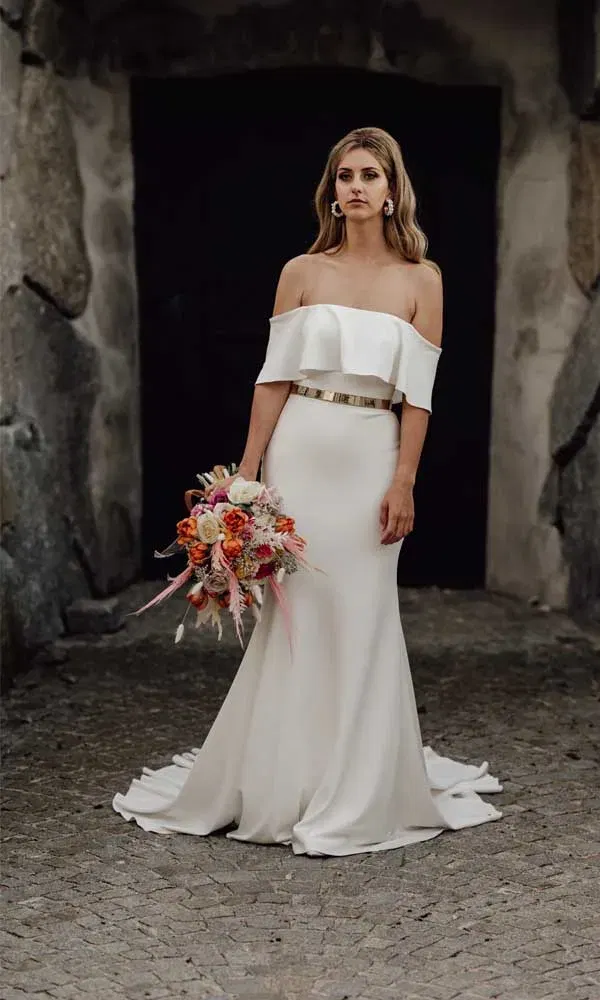 Schlichte Braut in weißem Off-Shoulder-Kleid mit goldfarbenem Gürtel, posiert mit Strauß vor dunklem Hintergrund