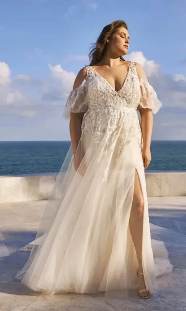 Eine kurvige Braut trägt ein elegantes Tüllkleid mit Schulterfreien Ärmeln und genießt die Meereskulisse im Sonnenlicht.