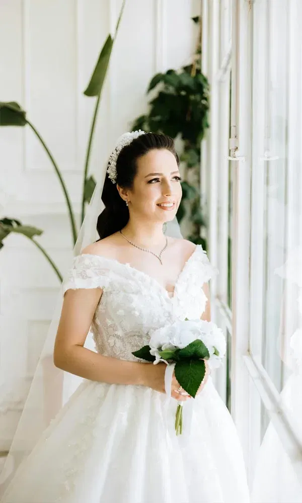 Junge Frau im Brautkleid steht mit Blumen in der Hand vor einem großen Fenster und strahlt.