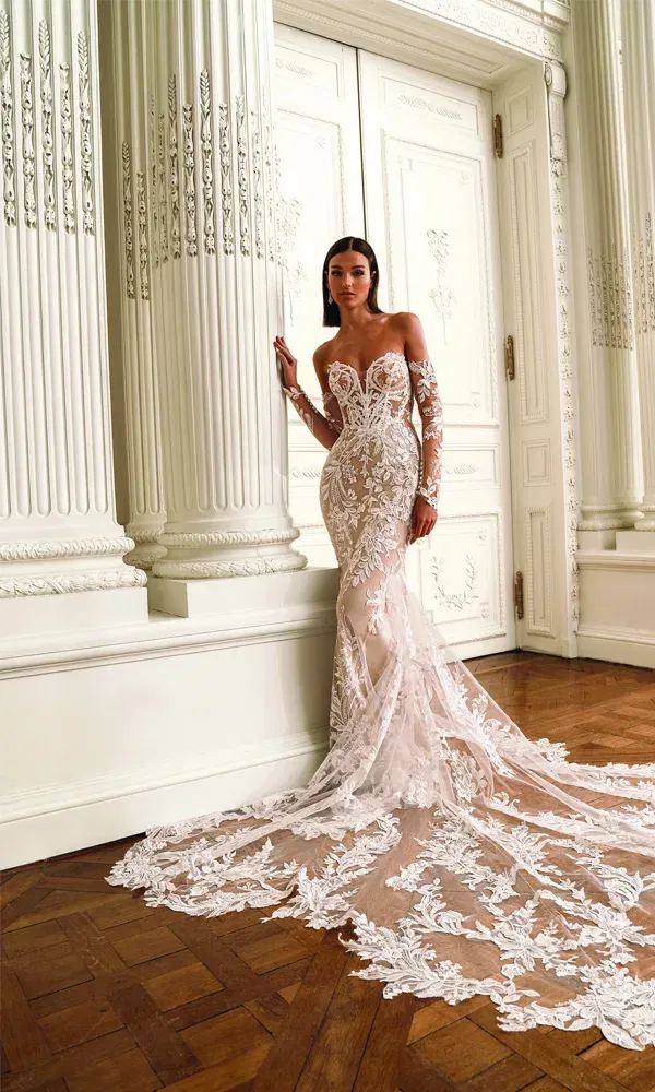 Elegante Braut in einem eng anliegenden, transparenten Hochzeitskleid mit floralen Stickereien und dramatischer Spitzenschleppe, stehend vor klassizistischer Architektur mit weißen Säulen.