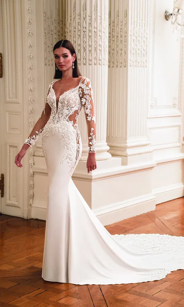 Model trägt ein figurbetontes Hochzeitskleid mit Stickereien und Illusionseinsätzen, posierend vor weißen Säulen und Wandverzierungen in einem herrschaftlichen Saal.