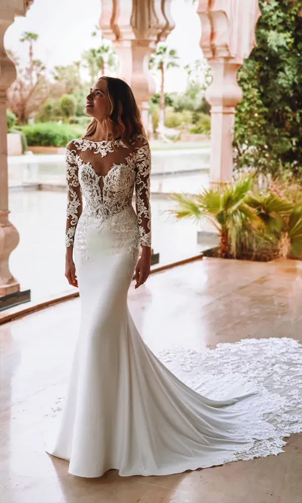 Model in einem Fit-and-Flare-Brautkleid mit aufwendigem Spitzenmuster und langem Tüllschleier, posierend auf einer überdachten Terrasse mit exotischen Palmen im Hintergrund.