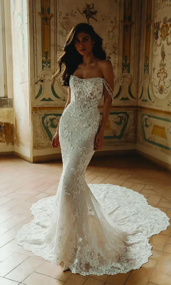 Elegante Braut in einem eng anliegenden Spitzenkleid mit zarten Schulterketten, posierend vor einer kunstvoll verzierten Wand in einem antiken Saal mit warmem Licht.