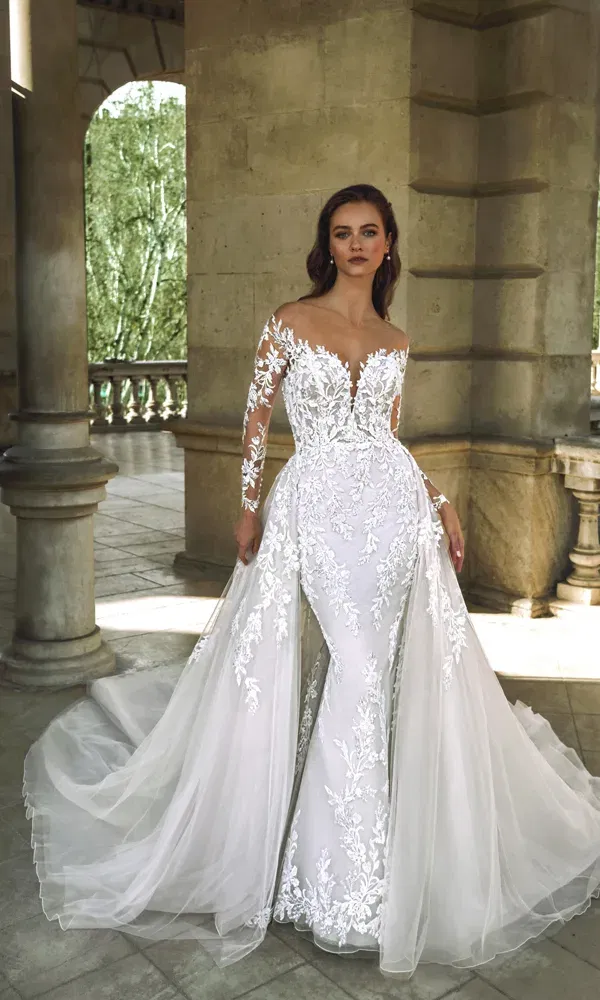 Elegante Braut in einem figurbetonten Hochzeitskleid mit langen Spitzenärmeln und voluminösem Overskirt, stehend vor klassischer Steinarchitektur mit Blick ins Grüne.