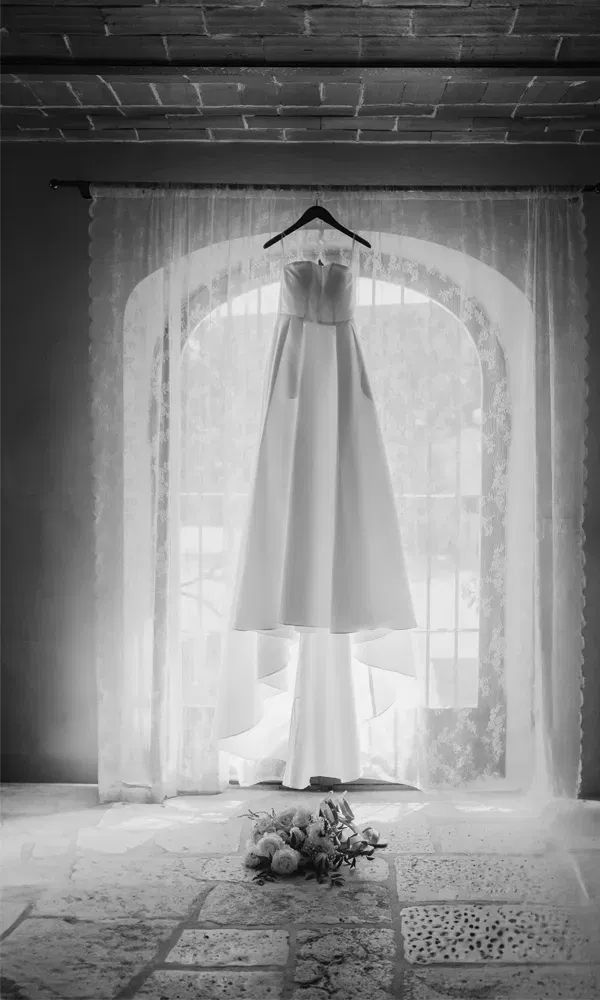 Hochzeitskleid auf einem Bügel vor einem großen Bogenfenster mit Lichtspiel – Aufnahme in Schwarz-Weiß mit romantischem Flair.