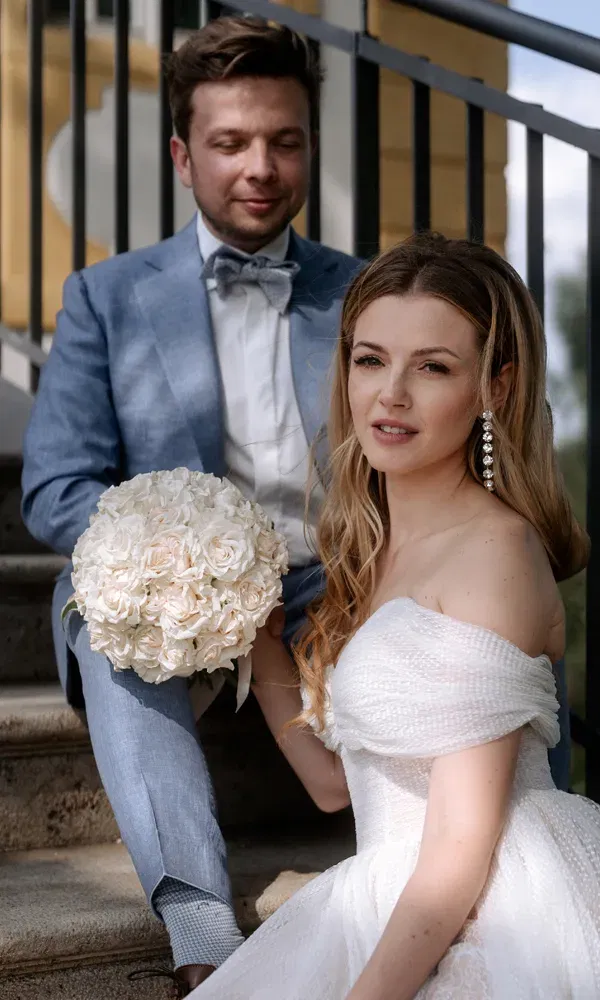Romantische Szene auf einer Steintreppe: Die Braut blickt in die Kamera, während der Bräutigam mit Blumen an ihrer Seite sitzt.