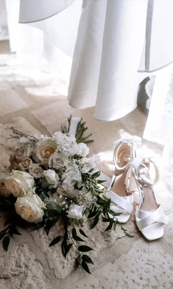 Detailaufnahme von Brautschuhen mit Schleifen und Blumenbouquet auf hellem Steinboden – zarter Moment vor der Trauung.