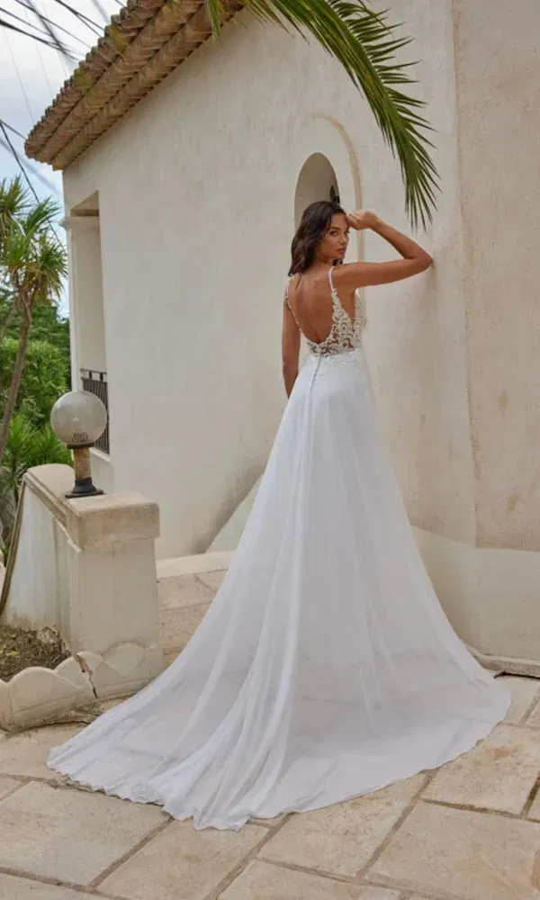Rückansicht einer Braut in einem fließenden Brautkleid mit Spitzenrücken und Spaghettiträgern auf sonniger Terrasse.