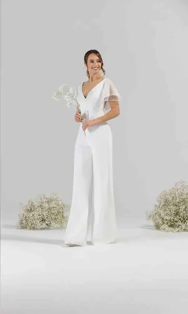 Lächelnde Frau im eleganten Hochzeits-Jumpsuit mit V-Ausschnitt und feinen Ärmeln vor minimalistischem Hintergrund.