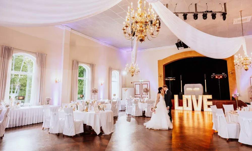 Romantischer Hochzeitssaal mit Kronleuchtern, weißen Stuhlhussen, stilvoller Deko und tanzendem Brautpaar vor leuchtendem LOVE-Zeichen.