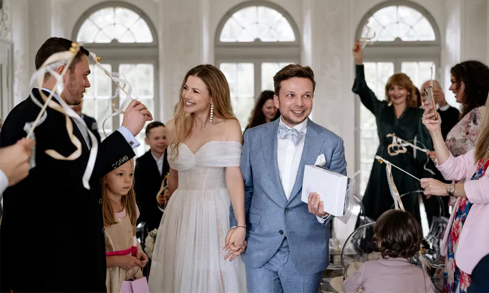 Strahlende Braut und Bräutigam betreten gemeinsam die Hochzeitslocation, während Freunde und Familie begeistert mit Bändern winken.