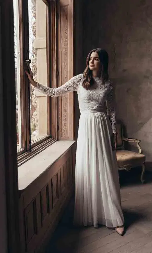 Zeitlose Brautszene mit einer Frau in einem zarten, langärmeligen Spitzenkleid vor einem historischen Fensterrahmen.