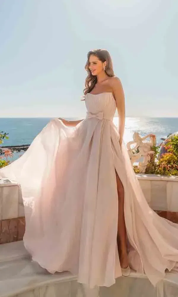Moderne Braut in fließendem Chiffonkleid mit hohem Beinschlitz posiert am sonnigen Meer auf Marmortreppen.