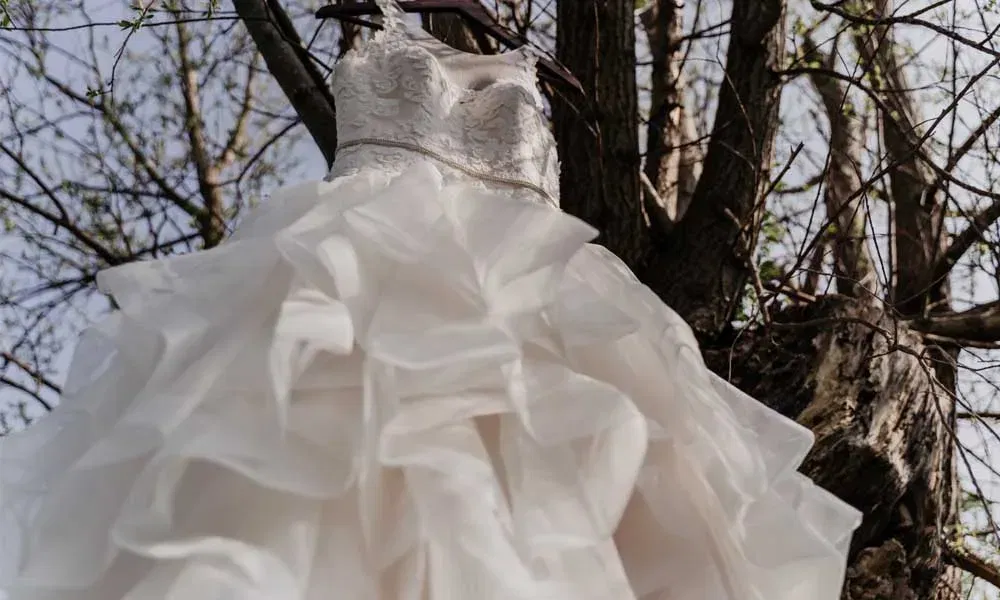Voluminöses Hochzeitskleid mit Spitzenoberteil und Lagenrock hängt an einem Baumstamm im Freien