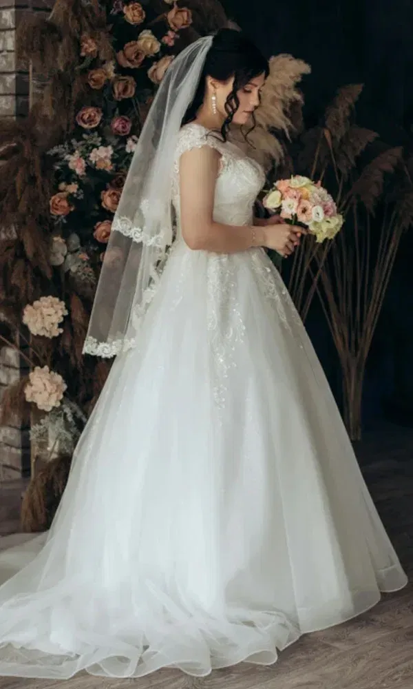 Frau im Brautkleid mit Spitzendetails und langem Schleier steht seitlich mit floraler Deko im Hintergrund.