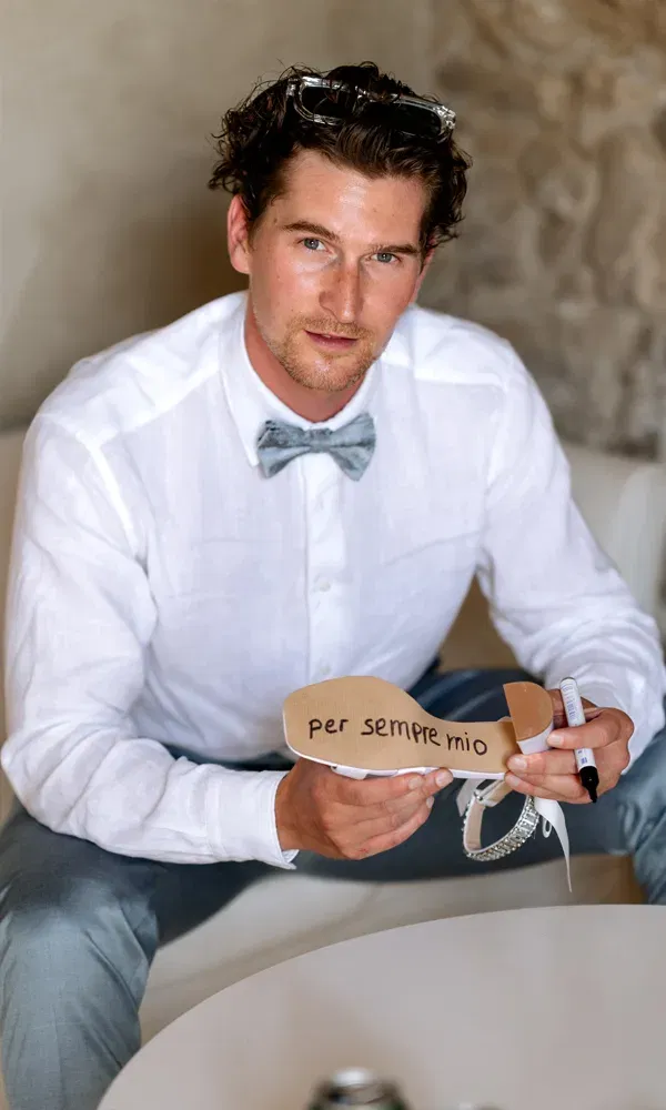 Lächelnder Bräutigam sitzt entspannt mit einem beschrifteten Schuh in der Hand – eine liebevolle Botschaft für seine Braut.