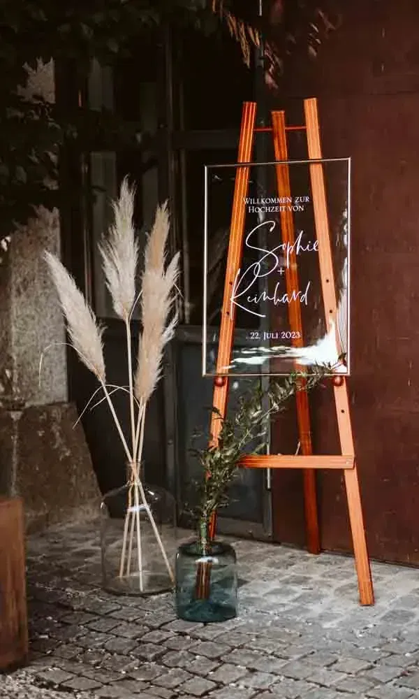Moderne Hochzeitstafel mit Schriftzug „Willkommen zur Hochzeit von Sophie & Reinhard“, umrahmt von Pampasgras und Olivenzweigen.