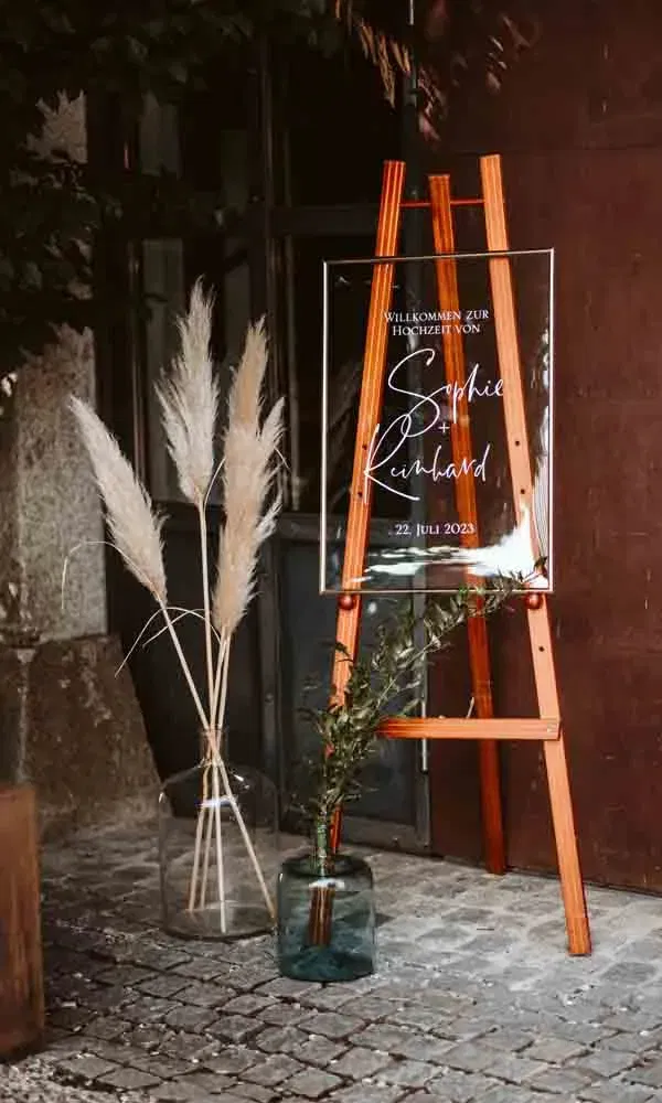Moderne Hochzeitstafel mit Schriftzug „Willkommen zur Hochzeit von Sophie & Reinhard“, umrahmt von Pampasgras und Olivenzweigen.